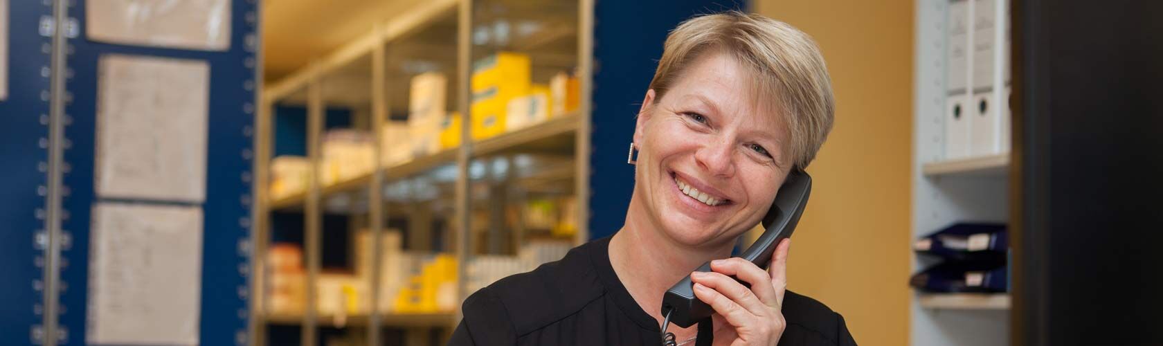 Eine lächelnde Frau telefoniert in einer Apotheke, umgeben von Regalen voller medizinischer Versorgung.