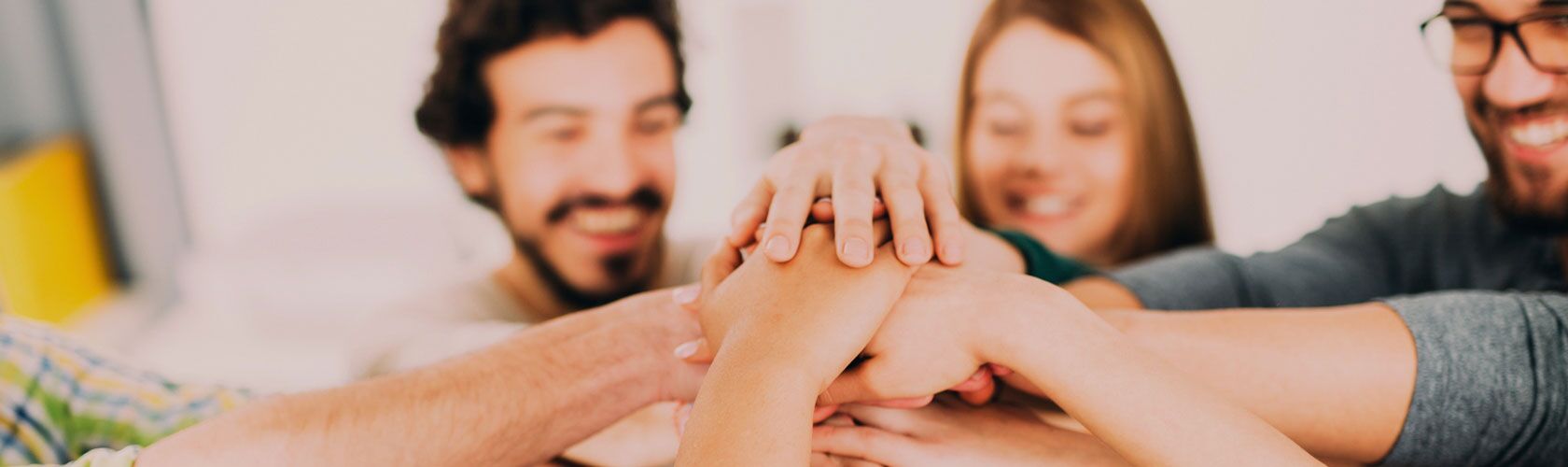 Ein Team von Personen legt lächelnd die Hände übereinander, um Zusammenarbeit und Willkommen zu symbolisieren.