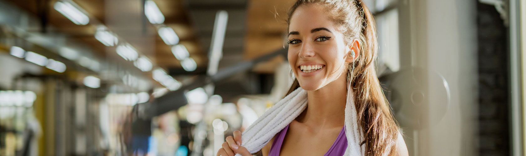 Eine lächelnde Frau im Fitnessstudio hält ein Handtuch um den Hals, was die Vorteile eines gesunden Lebensstils betont.