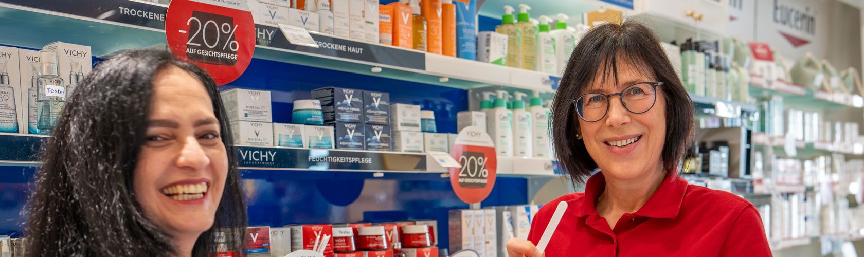 Zwei Frauen lächeln während einer Hautpflegeberatung vor einem Regal mit Dermo-Produkten in einer Apotheke.